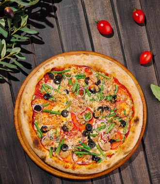 Sebze, zeytin ve otlarla kaplı taze pişmiş bir pizza ahşap bir masada oturur. Kiraz domatesleri ve zeytin dalları etrafa saçılmış, kırsal hissi arttırıyor..