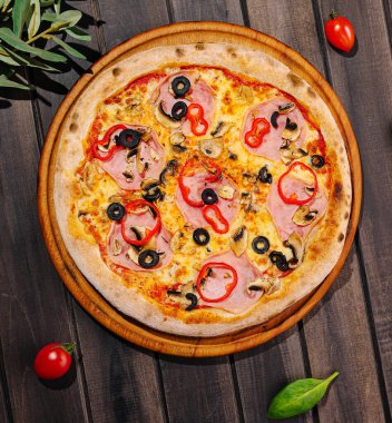 Bu lezzetli pizza çeşitli malzemeler içeriyor. Jambon, siyah zeytin ve çıtır çıtır sebzeler de dahil. Tahta servis tahtasında çok güzel sunuluyor..