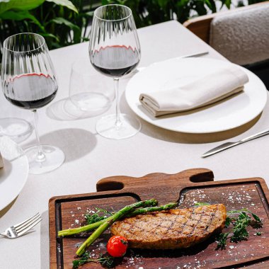 Izgara biftek, yanında kuşkonmaz ve vişneli domatesle ahşap bir tahtada sunulur. İki kadeh kırmızı şarap güzel bir sofraya oturup yemek deneyimini güçlendirir..