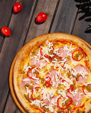 Yeni hazırlanmış bir pizza, üzerinde jambon, peynir ve renkli kırmızı biber bulunan ahşap bir tahtanın üzerinde oturur. Kiraz domatesleri etrafa saçılmış. Bu da canlı bir açık hava yemek deneyimi olduğunu gösteriyor..