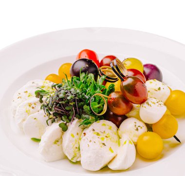 Canlı bir salata taze mozzarella topları, çeşitli renklerde üzümlü domatesler, ve beyaz bir tabakta enfes bir sunum sunan yeşillik ve otlardan oluşan bir garnitür içerir..