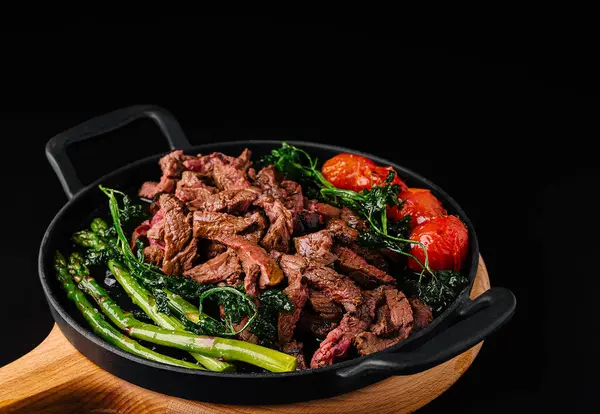 Lezzetli bir kızartmada taze sığır eti şeritleri parlak yeşil kuşkonmaz, yapraklı yeşiller ve olgun vişneli domatesler bulunur. Tabak basit bir tavada zengin renkler ve dokular sergiliyor.