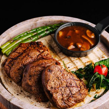 Lezzetli bir ızgara biftek, ahşap bir tahtada taze kuşkonmaz, otlar ve lezzetli bir sosla birlikte düzenlenir. Tabakta akşam yemeği için mükemmel bir yemek sergileniyor..