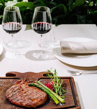 Güzel düzenlenmiş bir yemek masasında mükemmel ızgara bir biftek yanında kuşkonmaz ve süslemeler, yumuşak bitki aydınlatmasının altında iki bardak kırmızı şarapla eşleştirilmiş..