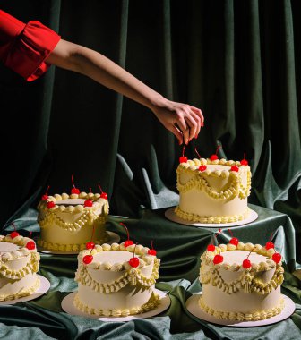 Bir el dikkatle birçok kremalı pastayı kirazlar ve zarif malzemelerle süsler. Ayar, neşeli etkinliğe şenlikli bir dokunuş ekleyerek, zengin yeşil kumaştan oluşuyor..