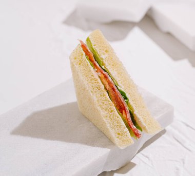 Lezzetli bir sandviç hafif bir yüzeyde dimdik durur, canlı sebzelerin katmanları sergilenir. Temiz sunumda taze malzemeler ve minimalist stil vurgulanıyor.