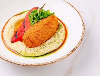 Sıcak bir kroket, yanında ızgara kırmızı biber ve taze kişniş ile süslenmiş kremalı humusun üzerinde dinlenir. Yemek canlı renklerle dolu, iştah açıcı dokuları vurguluyor..