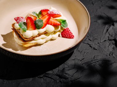 Çıtır kraker ve kremalı tatlı, taze çilek ve nane yapraklarıyla süslenir. Koyu, desenli bir yüzeyde güzelce sunulur..