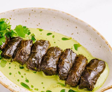 Lezzetli malzemelerle dolu üzüm yapraklı rulo bir tabakta zarif bir şekilde düzenlenir. Canlı bir yeşil sos tabağı çevreleyerek lezzetleri ve sunumu artırır. Taze bitkiler biraz renk katar..