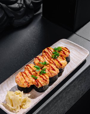 Izgara deniz ürünleri ile süslenmiş taze otlarla süslenmiş suşi rulolarının tadını çıkarmak. Havalı bir tabakta sunulan, turşulu zencefil ile tamamlanmış mükemmel bir yemek..