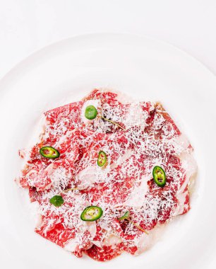 İnce dilimlenmiş et carpaccio, taze otlar, yeşil biber dilimleri ve rendelenmiş peynir tozuyla süslenmiş, canlı renkler ve dokular sergilenen beyaz bir tabakta güzelce düzenlenmiştir..