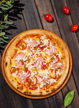 Yeni pişmiş bir pizza ahşap bir tabakta sunulur, jambon, kırmızı biber ve mozzarella peyniri ile süslenir. Kiraz domatesleri etrafa saçılmış, kırsal ayara renk katıyor..