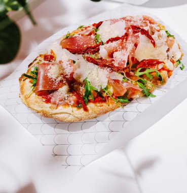 Prosciutto, roka, vişneli domates ve üzerine peynir serpiştirilmiş ağız sulandıran yassı ekmek, rahat bir yemek ortamında beyaz bir kağıtta oturur..