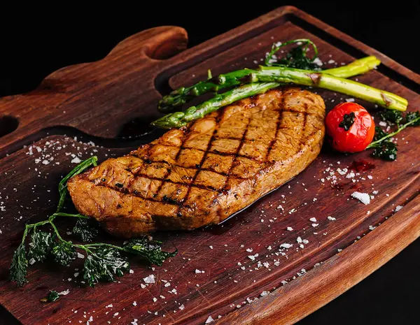 Mükemmel ızgara biftek ahşap bir tahtanın üzerinde duruyor, kuşkonmaz ve kiraz domatesleriyle süslenmiş. Tuz kristalleri bu iştah açıcı yemeğe biraz zarafet katar, doyurucu bir yemek için ideal..