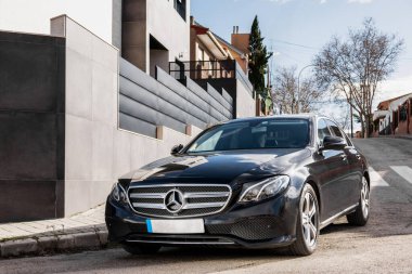 Parlak plakalı, parlak siyah bir Mercedes Benz yolun kenarına park edilmiş, lastikleri otomotiv aydınlatmasının altındaki kaldırımda duruyor.