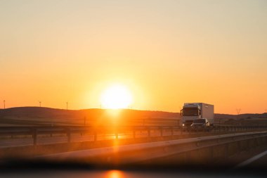 Bir kamyon gün batımında otomotiv ışıkları açık, gün batımından gün doğumuna kadar güneş ışığı yansıyan bir otoyolda gidiyor.