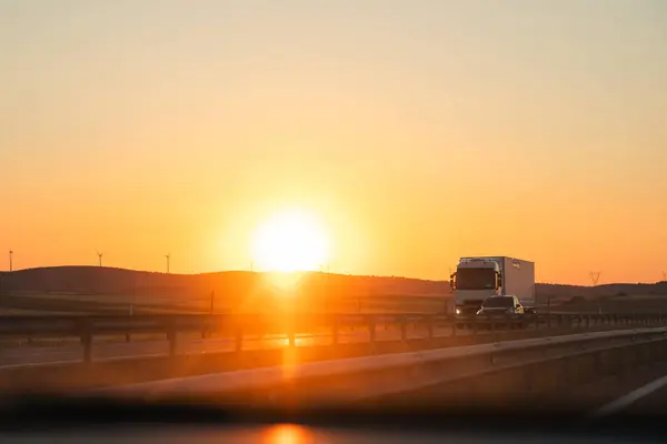 A truck drives on a highway at sunset, under the afterglow with automotive lighting on, sunlight reflecting on the water, and the sky transitioning from dusk to sunrise