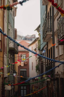 Bayraklar ve çiçeklerle süslenmiş iki bina arasında dar bir ara sokak, şehir manzarasında renk ve canlılığın bir karışımını sergiliyor.