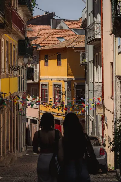 stock image Two women are strolling along a narrow street that is flanked by two buildings, under the open sky with windows and fixtures. The road surface is wellmaintained in the city