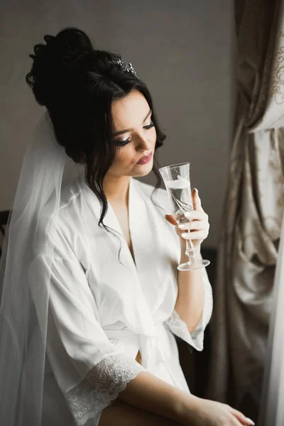 Beautiful and lovely bride in night gown and veil sitting and drinking champagne.