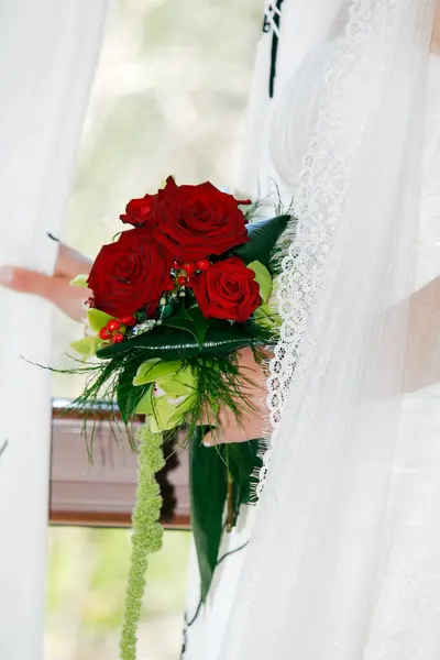 Bridal hands with bouquet of red roses.A hand of the bride holds the bouquet of flowers as she looks out the window on her wedding day.
