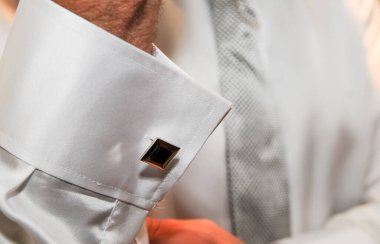 Bir adamın beyaz gömleğinin manşetini taktığı güzel bir kol düğmesinin yakın çekimi. Gömleğin manşetinde siyah ve gümüş süsler var..