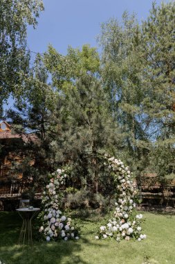 Serin bir bahçede sergilenen çarpıcı bir çiçek kemeri ve beyaz sandalyeli açık hava düğün töreni.