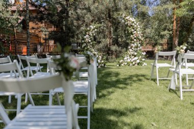 Serin bir bahçede sergilenen çarpıcı bir çiçek kemeri ve beyaz sandalyeli bir açık hava düğün töreni.