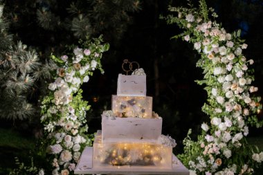 Romantik ışıklarla aydınlatılmış şık katlı bir düğün pastası, çiçeklerle süslenmiş bir arka planda.