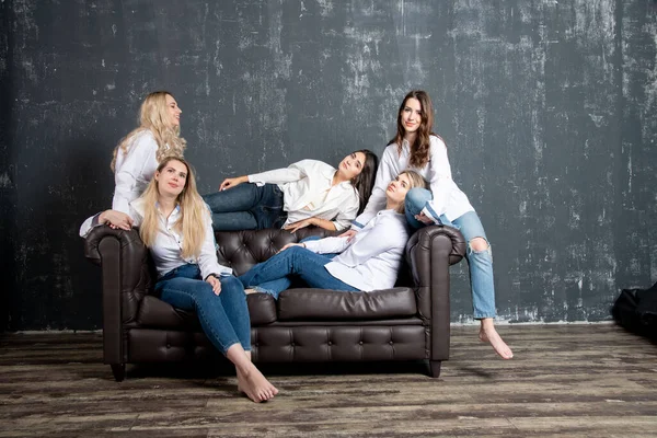 Group portrait of five attractive Caucasian women wearing white shirts and jeans posing on sofa.