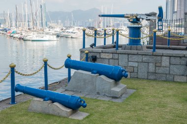 Two blue cannons sit on granite bases near Victoria Harbor, next to the Jardine Noonday Gun in Hong Kong. A chain link fence separates the cannons from the harbor. clipart