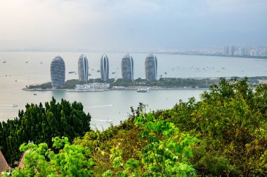 Sanya, Hainan, Çin 'deki Phoenix Adası' nda beş uzun ve ince binayla kıyı şehrine bakan panoramik bir manzara. Suda tekneler ve gemiler var.