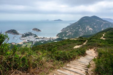 Dragon Back Hike, Shek O Peak, Hong Kong: Yeşil bir dağ yamacındaki dolambaçlı toprak yol kayalık burunların arasına kurulmuş bir kıyı köyüne bakıyor.