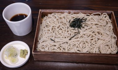 Tahta bir kutuda, deniz yosunlarıyla süslenmiş açık kahverengi soba erişteleri bulunur. Dilimlenmiş taze soğan ve wasabi içeren küçük bir tabak.. 