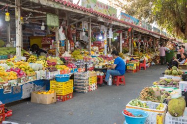  Sanya, Hainan, Çin - 19 Şubat 2024: Sanya 'da müşterilerin göz gezdirdiği ve satın aldığı hareketli bir meyve pazarı. Bir adam kırmızı bir taburede oturmuş, başka bir kadın telefonunu kullanırken meyve hazırlıyor..