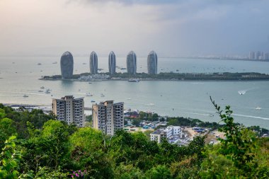 Sanya, Hainan, Çin 'deki Phoenix Adası' nda beş uzun ve ince binayla kıyı şehrine bakan panoramik bir manzara. Suda tekneler ve gemiler var.