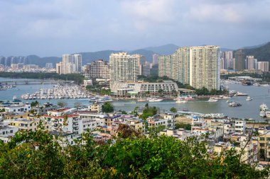 Sanya, Hainan, Çin 'in panoramik manzarası yüksek binalar, teknelerle dolu hareketli bir marina ve yemyeşil bir ön plan. Şehir manzarası arkadaki dağlara doğru uzanıyor..