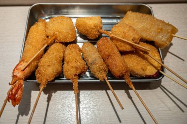 Bir metal tepside çeşitli kushiage karidesleri, sebzeler ve etler bulunur, hepsi bol yağda kızartılmış ve altın sarısı kahverengiler, şişler üzerinde servis edilir..