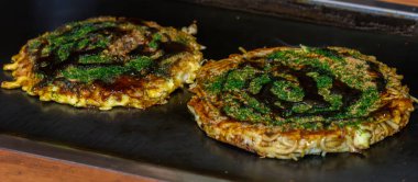 Iki okonomiyaki yakın çekim, lezzetli Japon gözlemeleri, sıcak bir ızgarada pişiyor. Üstünde aoknori yosunları ve okonomi sosu var..