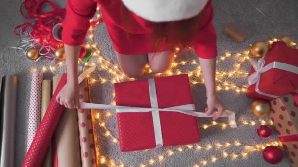 若い女性が友人や親戚のためにクリスマスプレゼントを梱包しています 女の子は赤い紙 色のリボンで新年のパッケージを作成します クリスマスの装飾 ホリデーシーズンの準備 — ストック動画