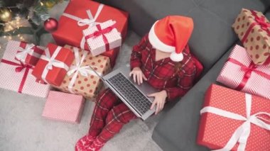 Noel Baba şapkalı bir kız ailesi ve arkadaşlarıyla bilgisayardan iletişim kuruyor. Evdeki kanepede dizüstü bilgisayarı, Noel ağacı ve hediyeleri olan kırmızı elbiseli bir kadın. Yeni yıl tatili ve alışveriş, internetten sipariş vermek..