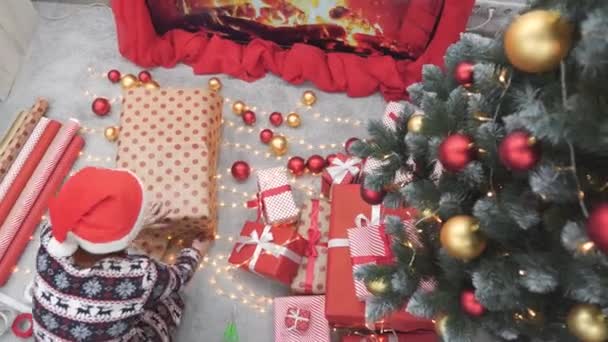 クリスマスツリーの近くのクリスマスライトと部屋の暖炉を背景に 多くの美しいクリスマスギフトボックスの近くの若い女性 雰囲気と気分新年 金と赤のお祝いの装飾 — ストック動画