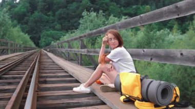 Dişi turist bir köprü üzerinde sırt çantasıyla oturur ve bekler. Turuncu sırt çantalı, şapkalı ve kameralı bir kız. Aktif ve turist yaşam tarzı kavramı.