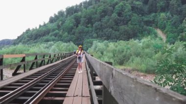 Bir kadın turist, köprüdeki tren yolu boyunca sırt çantasıyla yürüyor. Turuncu sırt çantalı, şapkalı ve kameralı bir kız. Aktif ve turist yaşam tarzı kavramı.