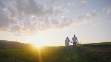 Mutlu çift sahada günbatımının arka planında eğleniyor. Birbirine aşık bir erkek ve kız yazın doğada yürüyor, sarılıyor ve dinleniyorlar. El ele tutuşup, gün batımını seyrediyorum..