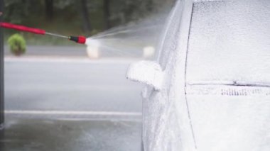 Bir adam oto yıkamada araba yıkar. Genç bir adam arabayı basınç altında köpük ve suyla temizler ve bir bezle siler. Gri bir arabayı özen ve yaz elle yıkama.