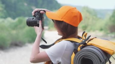 Bir turist kız gezi sırasında doğanın fotoğraflarını çeker. Kamera, turuncu sırt çantası ve şapkayla yürüyüş. Genç bir kadın DSLR kamerasıyla fotoğraf çekiyor. Ormanda ve dağlarda dinlenmek için bir yolculuk..