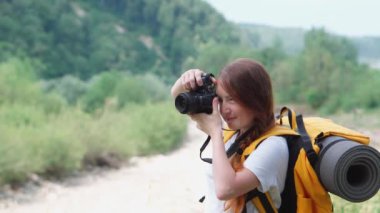 Bir turist kız gezi sırasında doğanın fotoğraflarını çeker. Kamera, turuncu sırt çantası ve şapkayla yürüyüş. Genç bir kadın DSLR kamerasıyla fotoğraf çekiyor. Ormanda ve dağlarda dinlenmek için bir yolculuk..