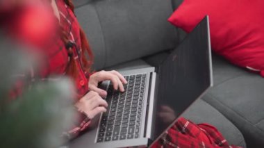 Evdeki kanepede dizüstü bilgisayarı, Noel ağacı ve hediyeleri olan kırmızı elbiseli bir kadın. Noel Baba şapkalı bir kız bilgisayarda video aracılığıyla iletişim kuruyor. İnternetten öğreniyorum. Yeni yıl alışverişi ve siparişi
