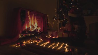 Arka planda Noel ışıkları, çelenkler ve şöminenin üzerinde Noel hediyesi kutuları olan bir kız. Genç bir kadın yeni yıl arifesinde hediyeleri ağacın yanında renkli kırmızı kağıtlarla sarar. Atmosfer havası, dekor kışı.